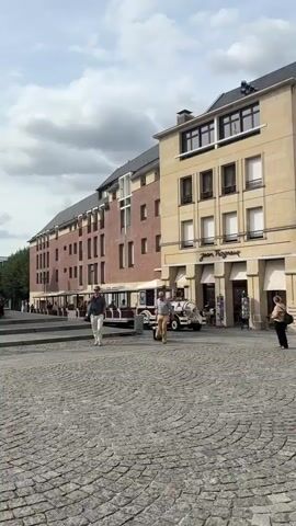 The video shows a street scene in a European city. The first shot is of a cobblestone plaza with people walking and a small train-like vehicle with passengers. The second shot focuses on a man walking with a bag, and the train with passengers continues in the background. The third shot shows a family sitting on steps in front of a historic building with a prominent tower. The fourth shot pans across a street with several buildings, including shops, and a few people walking. The final shot is a close-up of the facade of a large, ornate cathedral, with people walking up its steps. The overall environment appears to be a sunny day with some clouds.