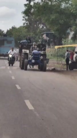 A blue and white tractor is driving down a paved road. A man is driving the tractor, wearing a checkered shirt and a scarf. The tractor is pulling a trailer with equipment on it. A motorcycle is driving on the left side of the tractor, and another tractor is visible in the background. There are trees and buildings on the side of the road. The sky is overcast.