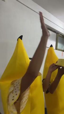 Two people in banana costumes are in a room. One person is on the left and is blurry. The person on the right is in the foreground and is also blurry. A woman in a yellow dress is standing to the right of the person in the foreground. There are stacks of boxes in the background. A man in a blue hat and plaid shirt is standing to the left of the person in the foreground. The lighting is dim.