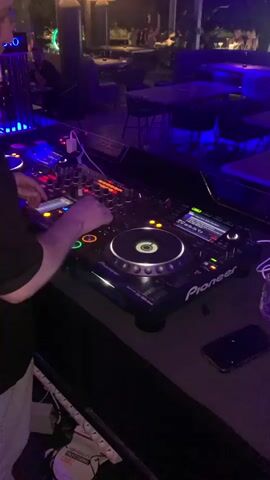 The video shows a DJ performing at a club. The DJ is standing behind a Pioneer DJ setup, which includes two CDJs and a mixer. The DJ's hands are on the equipment, and they appear to be adjusting the controls. The club is dimly lit, with blue and purple lights illuminating the scene. In the background, there are tables and chairs, and some people are seated. The DJ is wearing a dark shirt and light-colored pants. The overall atmosphere suggests a lively and energetic environment.
