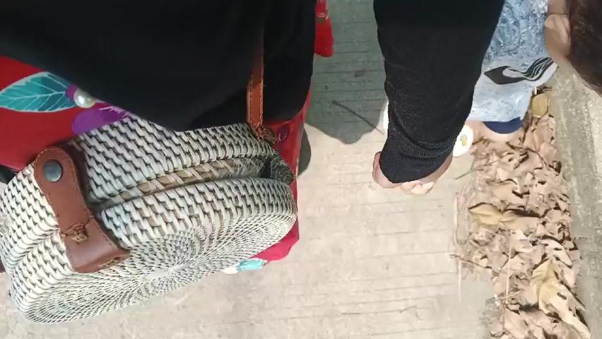 The video is filmed from a high-angle, over-the-shoulder perspective, looking down at the feet of two individuals. The left side of the frame shows a woven, round straw bag with leather straps and metal accents. A person's legs are visible, wearing a red dress with a floral pattern and light brown crocs. To the right of the bag, a child's legs are visible wearing grey socks with a black logo and white sandals with cartoon characters. The child is holding the hand of an adult, who is also wearing light brown crocs. The ground is a concrete pathway with dry leaves scattered on the right side. The scene appears to be outdoors in daylight. The action captured is the adult and child holding hands, seemingly walking or standing on the pathway.