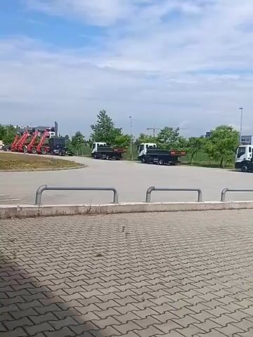 The video shows a parking lot with several trucks and a car. In the foreground, a blue car is parked facing right. Behind the car, there are multiple large, dark-colored dump trucks parked in a row, facing left. Some of these trucks have "Schwarzmüller" written on their sides. Further in the background, there are white trucks with flatbeds and a red piece of machinery, possibly a loader, on the far left. The parking lot is paved with interlocking concrete blocks. Trees and some buildings are visible in the distance under a partly cloudy sky. The camera appears to be moving slightly from left to right, panning across the scene.