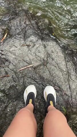 The video shows a person standing on a gray rock next to a body of water. The person is wearing white and black water shoes. The water is flowing into a pool, creating a waterfall. The rocks are dark and wet. There are some fallen leaves and small twigs on the rock surface. The sound of the flowing water is audible.