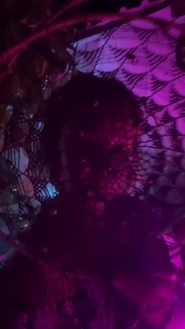 A person is captured in a dimly lit, vibrant setting, likely a nighttime party or festival in Arambol, India. Their face is largely obscured by a large, circular, net-like headpiece that appears to be intricately woven and adorned with small, reflective elements. Dark, curly hair is visible around the edges of the headpiece. The scene is illuminated by striking neon lighting, predominantly in shades of purple and blue, casting a dynamic and atmospheric glow. Hints of string lights and other festive decorations can be seen in the blurry background. The camera moves slightly, giving a dynamic perspective of the person amidst the lively ambiance. No visible text is present.