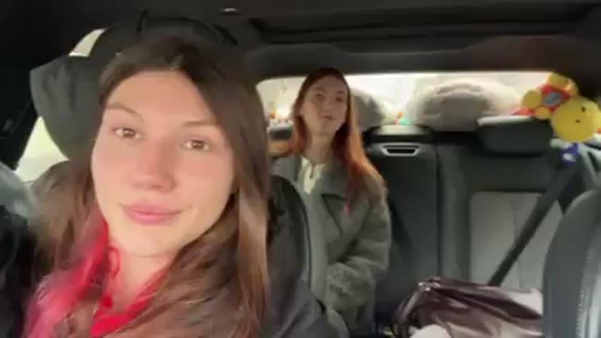 Two women are in the back seat of a car. The woman closest to the camera has dark hair with pink tips, and is wearing a black jacket with a red shirt underneath. She is smiling slightly. The woman in the back is a redhead, wearing a light grey fur-like jacket. She looks at the camera briefly. Between the women, a brown purse sits on the seat, and behind them are a couple of stuffed toys. The car appears to be a luxury vehicle with a sunroof and leather seats. Music plays in the background.