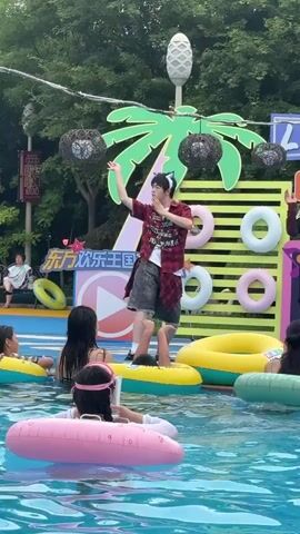 The video shows a young person wearing a red plaid shirt over a white t-shirt and dark shorts, standing on a platform in a swimming pool. They are wearing cat ears and making hand gestures. Several children are in the pool, some with inflatable rings. A backdrop behind the person features a large palm tree illustration and a sign with Chinese characters. There are also decorative hanging lights and inflatable rings on display. The environment appears to be an outdoor pool area.