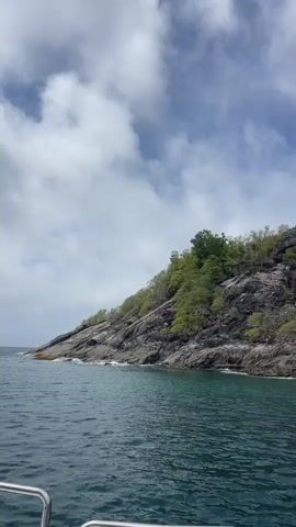 The video shows a boat moving through turquoise water towards a lush green island with rocky outcrops. The sky is cloudy with patches of blue. The boat appears to be a small vessel with a metal railing visible in the foreground. The island's coastline is characterized by dense vegetation and exposed rock faces, with waves gently lapping at the shore. The perspective suggests a slow, steady movement of the boat along the coast.