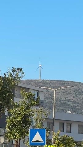 The video shows a stationary shot of a wind turbine spinning on a hill against a clear blue sky. A building, street lamp, foliage and a blue traffic sign are visible in the foreground. The wind turbine is white and has three blades. The hill is covered with sparse vegetation.