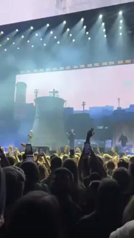 A low-angle shot shows a concert performance. A crowd of people faces a stage, holding up their phones to record. On the stage, three performers stand in front of a large screen displaying a backdrop resembling an industrial landscape, complete with smokestacks and dilapidated structures. The lighting on stage is bright, with numerous spotlights directed toward the performers. Fog or smoke effects enhance the atmosphere.