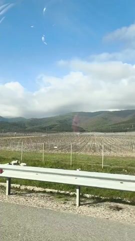 The video shows a view from a moving vehicle, likely a car, traveling along a road. A white guardrail runs parallel to the road. Beyond the guardrail, there is a grassy area. In the distance, there are trees, and in the far background, there are hills covered with some cloud cover. The sky is blue with patches of white clouds. The video appears to be shot during the day. The road surface is gray and shows signs of wear. At the start of the video, there's a fenced-in agricultural area, possibly a vineyard.