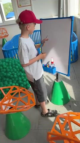 A child stands near a whiteboard in a room filled with colorful furniture and objects. The child is wearing a red baseball cap, a light grey T-shirt, dark grey shorts, and Crocs. The child is holding a marker and a brown eraser and writing on the whiteboard. The whiteboard is mounted on a stand with a blue frame and a green cone-shaped base. There is a blue container attached to the whiteboard stand, holding various markers and a spray bottle. To the left of the child is a ball pit filled with green balls. In the background are various colorful chairs and playpens. Natural light is streaming into the room through a window. The floor has a light-colored tile pattern.