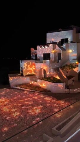 The video shows a three-story white building at night with a dynamic light projection. The projection includes abstract patterns and animated images, such as a glowing silhouette of a person reading a book. The light covers the walls of the building and spills onto the ground below. A small tree sits next to a staircase, and some short grasses are present in the landscaped areas. The background is dark with a suggestion of the sea visible on the horizon. The camera angle is a high vantage point.