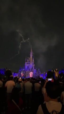 A crowd of people gathers in front of a large, illuminated castle at night. The castle is lit with blue and purple lights, and projections are visible on its facade. Two fireworks are exploding in the sky above the castle. Many people in the crowd are holding up their phones to record the spectacle. The sky is dark and cloudy.