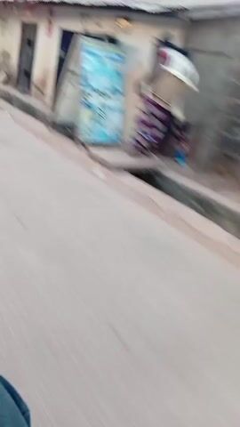 A person wearing jean shorts, a striped shirt, and sneakers walks along a street with low buildings on either side. A motorcycle is parked near the beginning of the clip. The camera appears to be moving forward, following the person walking. The scene is captured from a low angle, slightly angled downwards towards the street.