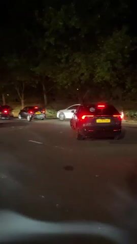 The video, shot at night, shows a parking lot with several cars driving and parked. The scene is illuminated by overhead streetlights, and trees surround the parking area. Initially, a cloud of vapor or smoke briefly obscures the foreground view before dissipating. Cars with illuminated red taillights move slowly in the parking lot. The camera pans slowly to show the extent of the parking area, revealing rows of parked cars and vehicles driving along the lanes.
