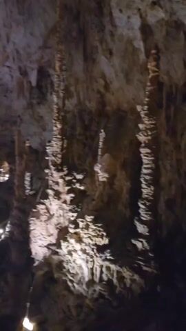 The video pans across a dimly lit cave interior filled with stalactites and stalagmites. Several large, columnar formations dominate the foreground and middle ground, illuminated by strategically placed lights that cast dramatic shadows. In the background, a wooden walkway with railings is visible, featuring small lights and a few indistinct figures of people walking along it. The cave walls and ceiling are rough and textured, showing layers of rock and mineral deposits. The overall impression is of a natural underground cavern that has been made accessible to visitors.