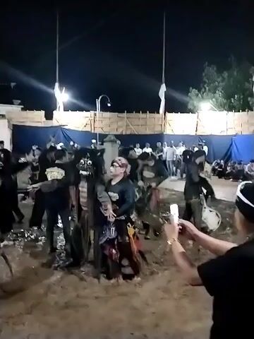 The video captures an outdoor night event in Jenggrik, Indonesia, featuring a group of men participating in what appears to be a traditional Indonesian dance or performance. The performers are dressed in dark attire, with some wearing elaborate costumes that resemble animal figures or traditional dance outfits. They are moving in a ritualistic or synchronized manner in an open area with a dirt ground.

Around the performance space, spectators are gathered, some seated and others standing, behind temporary bamboo fencing and blue tarpaulin barriers. Tall poles bearing white flags are also visible, illuminated by artificial lighting, suggesting the event is taking place after dark. In the immediate foreground, a person is actively filming the performance with a smartphone. The scene depicts a lively cultural gathering or community celebration. No distinct text is visible.
