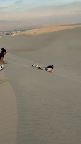 A person is lying prone on a sand dune, slowly sliding downhill. The person is wearing shorts and sneakers. A four-wheel drive vehicle with several people nearby is parked on another dune in the background. The environment is a desert landscape, with visible dunes, and a somewhat hazy sky. The dunes display tire tracks and footprints.
