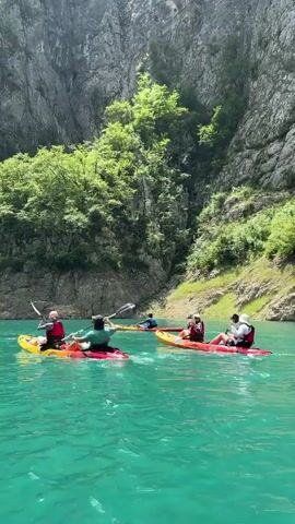 A group of people are kayaking on a turquoise lake. They are in a canyon with steep, rocky walls covered in trees. The sky is blue with some clouds. The people are wearing life vests and using paddles to move their kayaks through the water. One person in the front of the group is wearing a yellow kayak and a red life vest, and another person behind them is in a red kayak and a green life vest. There are other kayaks visible further back in the water, also with people wearing life vests. The water is calm and clear, and the sunlight is reflecting off its surface.