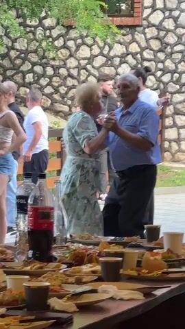 The video shows a gathering of people outdoors, possibly a party or celebration. In the foreground, a long, rustic wooden table is laden with plates of food, empty cups, and bottles of drinks, suggesting a meal has recently concluded or is ongoing. The table is messy, with remnants of food scattered across the surfaces. In the mid-ground, an elderly couple is dancing together, the man in a blue short-sleeved collared shirt and dark pants, and the woman in a light-colored floral dress. They are holding hands and appear to be in an embrace, looking at each other. Behind them, other individuals are visible. To the left, a young woman with long hair wearing a white top and denim shorts is partially visible, standing near a table with bottles. To the right and further back, a young man in a white long-sleeved shirt and dark pants stands facing the dancing couple. Another man in a black t-shirt is visible behind him. In the background, a stone wall forms the backdrop, with some greenery and trees above it. The lighting suggests it is daytime, possibly overcast. The overall atmosphere seems relaxed and festive.