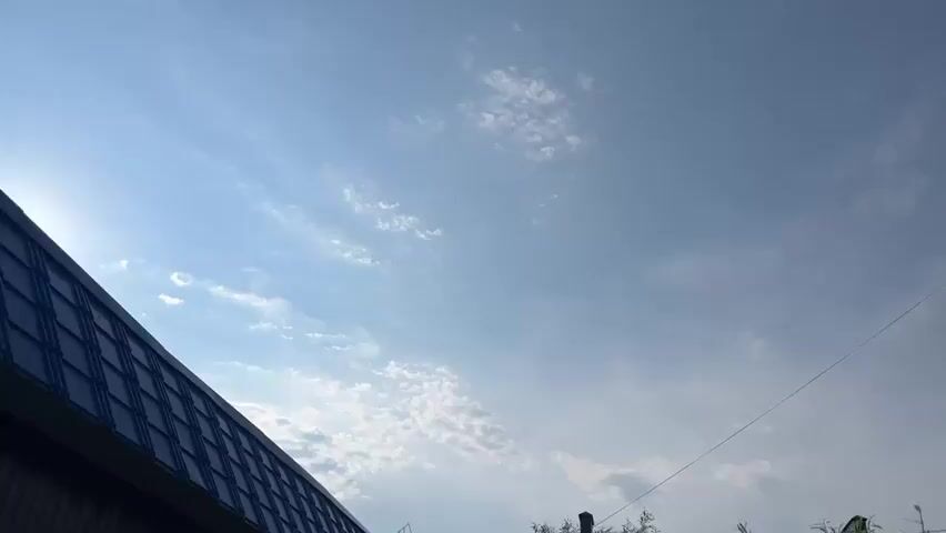 This is a landscape video with a focus on the sky and architecture. The scene transitions from a lower angle revealing the edge of a dark-tiled roof in the foreground on the left, to a higher angle showing the side of a modern building with a tiled facade in white, green, and orange squares on the right. A thin wire stretches diagonally across the frame, from the lower left towards the upper right.

The sky is a clear, pale blue with scattered white clouds. The time of day appears to be daytime, likely late morning or early afternoon, given the brightness of the sky and the angle of the implied sunlight. The weather seems fair.

The setting appears to be a residential or commercial area, possibly in Muravlenko, Russia, as indicated by the provided location context. The architecture suggests a contemporary construction. There are no people visible in the video, and the primary subjects are the sky and the buildings. The overall mood is calm and observational. There is no visible text within the video frame.