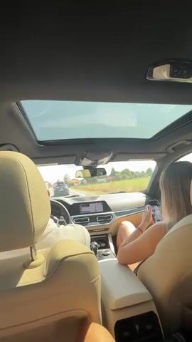 This is a first-person perspective video taken from the back seat of a car. The car has a sunroof open, and the sky is visible. The front passenger is looking at their phone. The driver is holding the steering wheel, and the front windshield shows a road with other cars and a rural landscape with fields and trees. The video pans to the right, showing more of the landscape through the side window. A person in the back seat is visible, with their legs extended. The interior of the car appears to be luxurious, with leather seats. The overall setting is a sunny day, likely a road trip through the countryside.