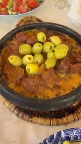 The video shows a close-up view of a tagine dish on a table. The tagine is a shallow, black, circular cooking pot filled with a stew-like mixture of meat and large green olives. The dish is steaming slightly, suggesting it is freshly cooked. The tagine pot sits on a woven, round trivet. To the left of the tagine is a plate of salad with chopped tomatoes and cucumbers. In the background, there's a glass and a plate with a blue and white floral pattern containing cucumber slices. The table is covered with a white tablecloth.