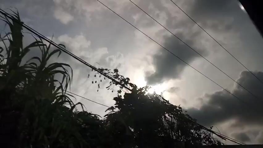 This is a low-angle, point-of-view video taken from a vehicle. The camera looks upward at a cloudy sky with the sun shining through. Several power lines are visible stretching across the frame. In the foreground, the dark silhouette of a building's roofline is visible on the left. As the vehicle moves, trees and two telephone poles appear and disappear. The overall impression is of a cloudy, possibly overcast day with intermittent sunlight.