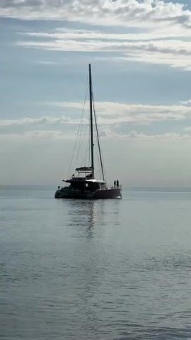 A serene scene unfolds with a large catamaran sailboat positioned in the calm, reflective waters of what appears to be a sea or ocean. The boat is silhouetted against a bright, cloud-dotted sky, suggesting either early morning or late afternoon. A few individuals can be seen standing on the stern of the catamaran, observing the surroundings. The water is smooth with minimal ripples, reflecting the soft light from the sky. The boat itself is a modern design with a tall mast, and its dark profile contrasts with the lighter hues of the sky and water. No text is visible in the video.