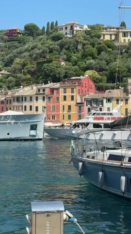 This is a short video clip showcasing the picturesque harbor of Portofino, Italy, on a bright, sunny day. The primary subjects are several yachts and boats docked in the clear blue water, with the colorful buildings of the town climbing up the lush, green hillside in the background.

In the foreground, a modern sailboat with a grey hull and white accents is prominently featured, tied to a stone quay. Its mast and rigging are visible against the sky, and it has several fenders hanging over its side. Further out in the harbor, a larger, white motor yacht is anchored, with people visible on its decks, suggesting a leisurely activity. Another large, light-colored vessel is docked on the left side of the frame. The distinctive, vibrantly colored buildings, characteristic of Portofino, are a focal point, with hues of pink, yellow, and orange dominating the scene. The hillside is densely covered with olive trees and other Mediterranean vegetation.

The activity taking place is primarily the docking and anchoring of boats in the harbor, implying a scene of leisure and tourism. The weather appears to be warm and clear, indicated by the bright sunlight and the deep blue sky. The time of day is likely midday, given the strong shadows and the intensity of the light. The overall emotion evoked is one of tranquility, beauty, and a sense of vacation or high-end travel.

There is some visible text on the hull of the sailboat in the foreground. It appears to read "AE 2824 D".