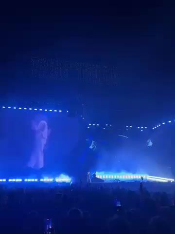 The video captures a live music concert at night, likely in a large outdoor venue or arena, consistent with a major festival. A performer, appearing to be a male in light-colored clothing, stands on a stage bathed in intense blue stage lights and spotlights. Behind him, a massive, irregularly shaped video screen displays a larger-than-life, semi-transparent image of a person, seemingly interacting with the live performance.

The foreground shows a large audience, their heads and numerous raised cell phones visible, indicating an engaged crowd capturing the event. The entire scene is dominated by the blue lighting from the stage, creating a vibrant and immersive atmosphere.

At approximately 0:02 into the video, large, white, illuminated text spelling "THERUPUTS" appears above the main screen and stage, serving as a prominent visual element. The setting suggests a large-scale event, possibly in Indio, United States, known for its music festivals. The overall mood is energetic and focused on the artistic spectacle presented on stage.