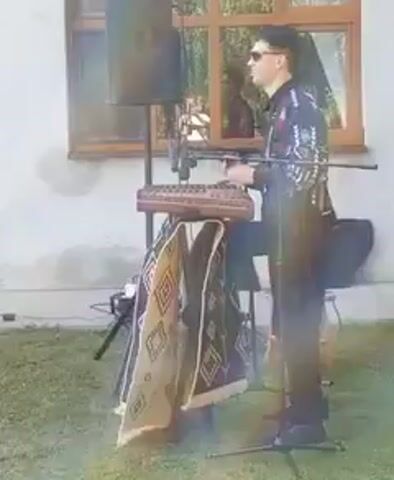 A man stands outdoors playing a dulcimer. He is wearing sunglasses and a black shirt with ornate white and red embellishments. The dulcimer is on a stand draped with a patterned rug. A speaker is behind him, and a microphone is in front of the instrument. The background shows a building with a window and trees.