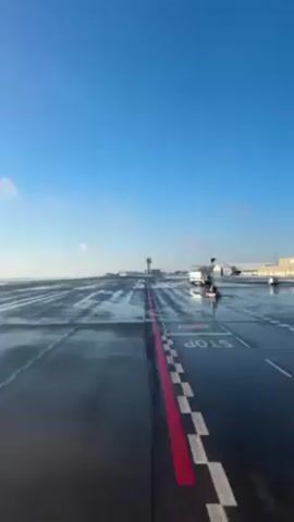 The video shows a wet airport runway on a sunny day with a clear blue sky. The runway is dark grey, with a red and white "STOP" line. In the distance, there is an air traffic control tower and other airport buildings. There are also vehicles on the runway. The camera appears to be moving slowly along the runway.