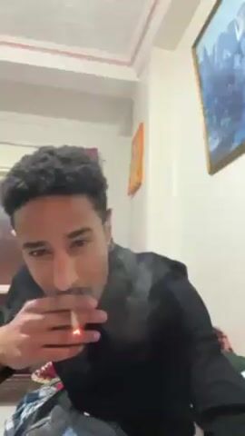 A young man with dark, curly hair and a mustache is smoking a cigarette in an indoor setting, facing the camera directly. He is wearing a black turtleneck and appears to be sitting, with bedding and clothing visible around him. The room is decorated with a picture hanging on the wall and has a simple ceiling with white tiles and pink trim. The man exhales smoke towards the camera, creating a visible cloud around his face. Music plays in the background.