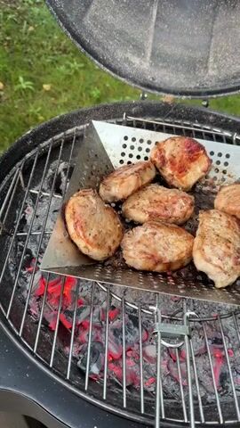 A top-down, close-up shot shows several pieces of pork chops being grilled over glowing red coals in a black charcoal grill. The pork chops are arranged on a metal grill basket that is placed on top of the main grill grate. The chops have a seared, browned exterior with visible seasoning. Smoke rises from the grill, partially obscuring the scene at times. The environment appears to be outdoors, with blurred green foliage visible in the background. The grill lid is partially open, allowing smoke to escape.