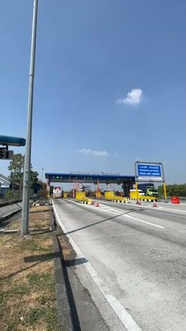 A clear, wide shot of a toll plaza on a sunny day. The toll plaza has multiple lanes, some of which are open with vehicles passing through. There is signage in Indonesian, indicating the toll plaza name, and a sign in English on the right listing special lane information. The sky is blue with a few scattered clouds. The road surface is asphalt, and there is grass and dirt on the sides. A tall metal pole stands on the left side of the frame.