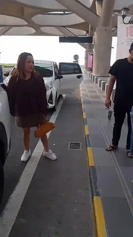 A woman in a brown top and short skirt exits a white car, holding a purse. A man in a black t-shirt and denim shorts loads luggage into the car. Another man in a blue shirt stands nearby. They appear to be at an airport or transportation hub, with architectural structures and signage visible in the background. The ground is paved with gray tiles.