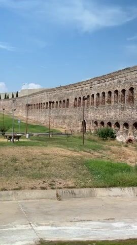The video pans across the exterior of a long, ancient stone wall with arched openings. The wall appears to be made of rough-hewn stones and brick. There are two distinct rows of arches. The upper row has larger, rectangular openings, while the lower row has smaller, rounded arches. Some of the upper openings reveal glimpses of trees and buildings in the distance. The wall is situated on a slight incline, with a grassy area in front of it. A concrete curb and a road are visible at the bottom of the frame. The sky above is blue with scattered clouds. Two light poles with visible fixtures are present in front of the wall. The lower portion of the scene shows a green, algae-covered body of water. The overall impression is one of historical ruins and a natural landscape.