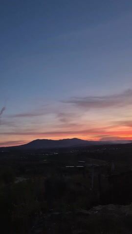 The video shows a sunset landscape. The sky transitions from deep blue at the top to orange and red hues near the horizon, with clouds streaking across the colorful portion. In the midground, there are silhouettes of mountains and a valley dotted with some lights, suggesting a settlement. The foreground is mostly dark, with the outlines of vegetation visible. Towards the end of the video, the camera pans slightly to reveal a portion of a white building with a tiled roof on the right side of the frame, with a small dish visible on a surface nearby.
