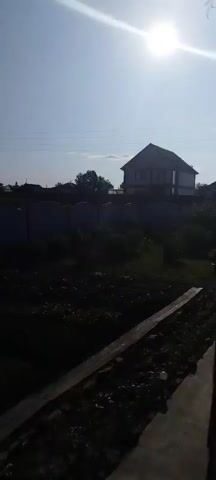 The video shows a garden with rows of plants. A white fence with a dark border runs across the middle ground, with a house and trees visible beyond it. The sky is clear and bright.