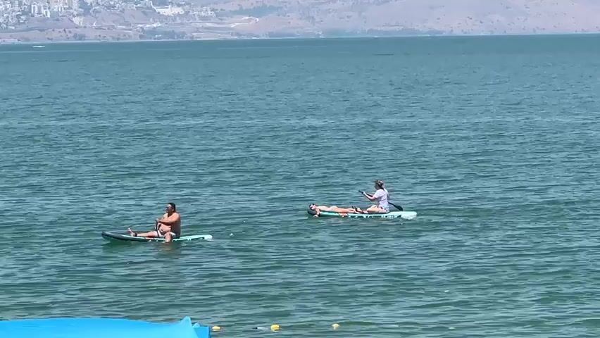 Two people are on paddleboards in a large body of water. The person on the left is a male wearing dark shorts and is paddling with a black paddle. The person on the right is a female wearing a white shirt and shorts, and is lying down on the paddleboard. The water is blue and calm, and there is land with buildings and hills in the background. The sky is overcast.