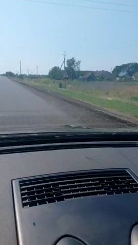 A first-person perspective shot from inside a car shows a road stretching ahead. The road is paved and has a white line marking the left lane. On either side of the road, there is vegetation and some houses are visible in the distance. The sky is clear and blue. Power lines run along the right side of the road. The dashboard of the car is partially visible in the foreground.