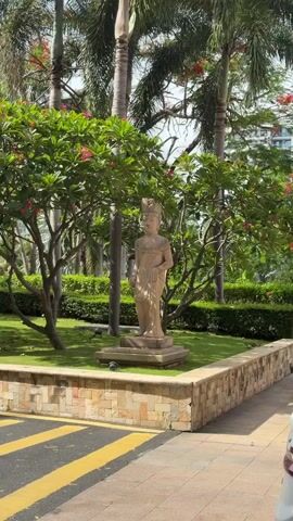 The video shows a serene garden with a stone statue of a person in the center. The statue is wearing a crown and is standing in front of lush green bushes and trees with red flowers. In the background, a tall, modern building stands against a bright sky. The foreground features a yellow and black striped pavement, and the top of a white car is visible on the right side. The overall atmosphere is peaceful and tropical.