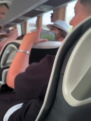 A low-angle shot from inside a bus shows several people, presumably passengers, seated in the vehicle. A person in the foreground, wearing a black shirt and a silver bracelet, holds a smartphone displaying a video of a soccer match, specifically, the English Premier League. A woman in a white hat, seated directly in front of the person holding the phone, is also watching the video on the phone. A second video showing a similar soccer match is on a screen mounted on the bus's ceiling. Other passengers can be seen, blurred, in the background of the bus. The video is a handheld shot and shaky.