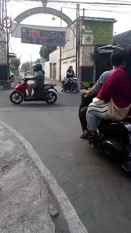 A motorcycle passes under an archway with banners. The motorcycle is driven by a person wearing a dark hijab and a red top, with a passenger in a grey top and white pants. Another motorcycle with a rider in a white shirt and dark pants passes from the opposite direction. In the background, other motorcycles are visible on the road, and buildings line the street. The ground is paved, with a cracked sidewalk in the foreground. The sky is overcast.