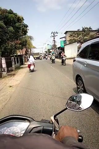 The video shows a first-person perspective of someone riding a motorcycle on a road. Several other motorcycles, some carrying one or two people, are also on the road. There are buildings and trees lining the road, and power lines are visible overhead. The sky is clear and blue. The rider's hand and a portion of the motorcycle's dashboard are visible in the foreground. The overall impression is a busy street scene in a populated area.