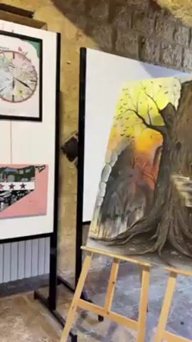 The video showcases an art exhibit displayed in a room with stone walls. The camera pans across various artworks, mainly depicting the theme of Syria.  There are several white display boards with framed and unframed artworks. Some of the art features the outline of the Syrian map, with various designs and collages inside. One piece is a clock-like diagram, and another is a painting of a tree against a sunset background on an easel. The lighting is dim, but highlights the art pieces.