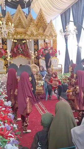 A wedding ceremony is taking place under a white canopy. The bride and groom are seated on a decorated stage, adorned in traditional attire. Several women in matching burgundy outfits and headscarves stand to the left of the bride, holding what appear to be fans. To the right, a group of women in similar attire are also present. In the foreground, several guests, including a child, are visible. The overall atmosphere suggests a festive cultural celebration.