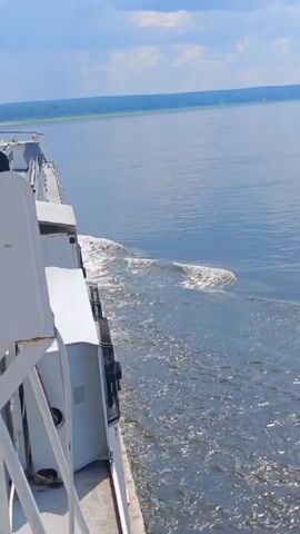 The video shows a side view of a boat moving through calm water. The boat is white with some grey sections. On the left side of the frame, part of the boat's superstructure is visible, including what appears to be equipment or storage units. The water is blue and reflects the sky, with a wake trailing behind the boat, indicating movement. The horizon in the distance is lined with green trees and a pale blue sky with scattered clouds. The camera appears to be positioned on the boat, looking backward and slightly to the side.