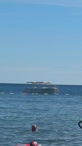 The video opens on a wide shot of the ocean under a clear blue sky. A small white boat with a canopy floats in the water. In the foreground, a person is swimming. Further in the foreground, a person is in a pink inflatable ring. The camera then pans to the beach. The beach is covered in dark pebbles and sand. Several beach towels are laid out on the ground, one with colorful stripes and another with a pink towel draped over it. Beach umbrellas and chairs are set up, and several people are relaxing on the beach. In the distance, more boats can be seen on the water, and a hilly coastline is visible on the horizon. The video ends with a close-up of two sets of bare feet resting on the pebble beach.